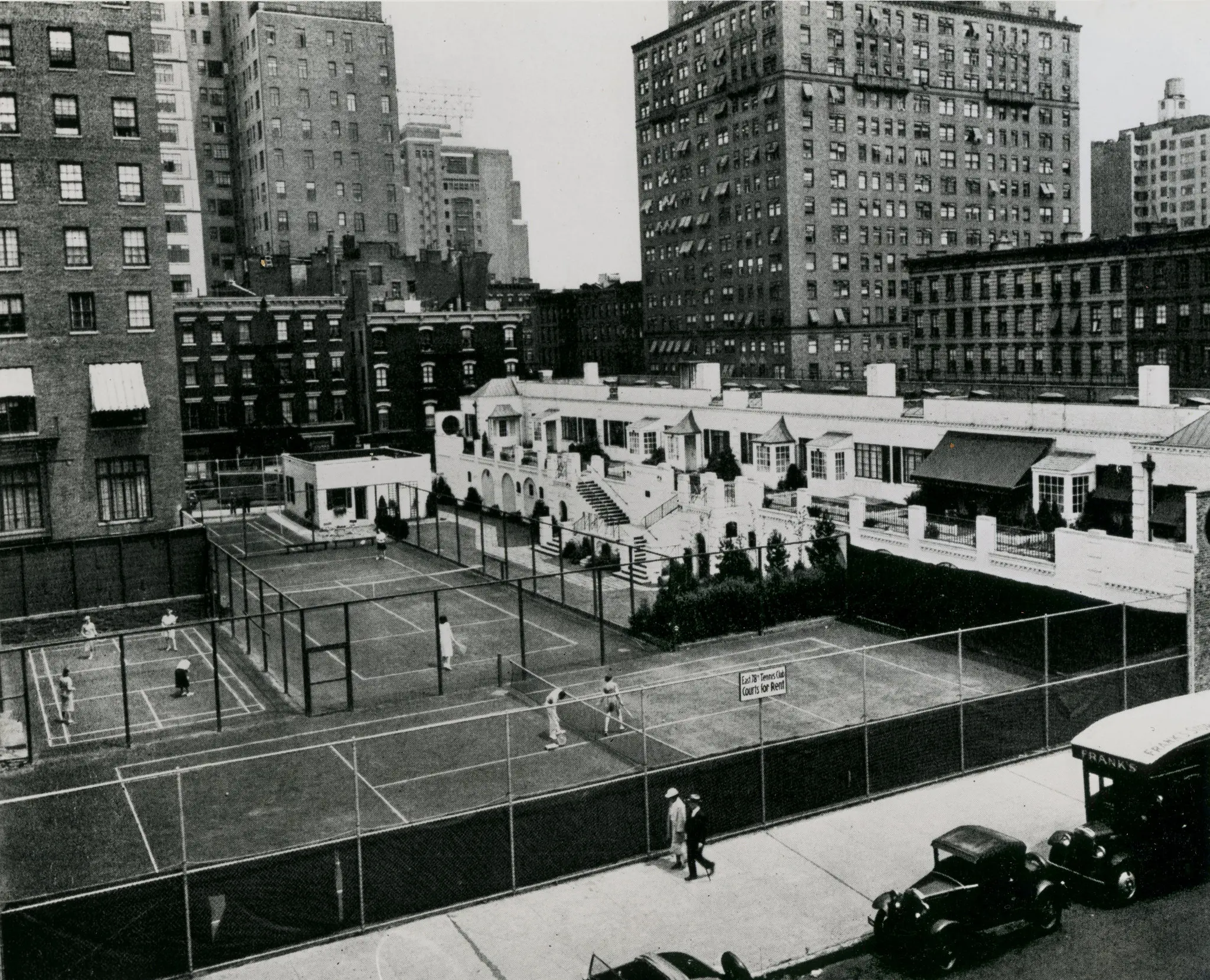 Photograph looking across two fenced tennis courts in the middle of a Manhattan block. Players in white are mid-game. Behind the courts stands a long, low, white building with a row of small projecting porches and a double staircase; tall brick apartment houses rise behind it.
