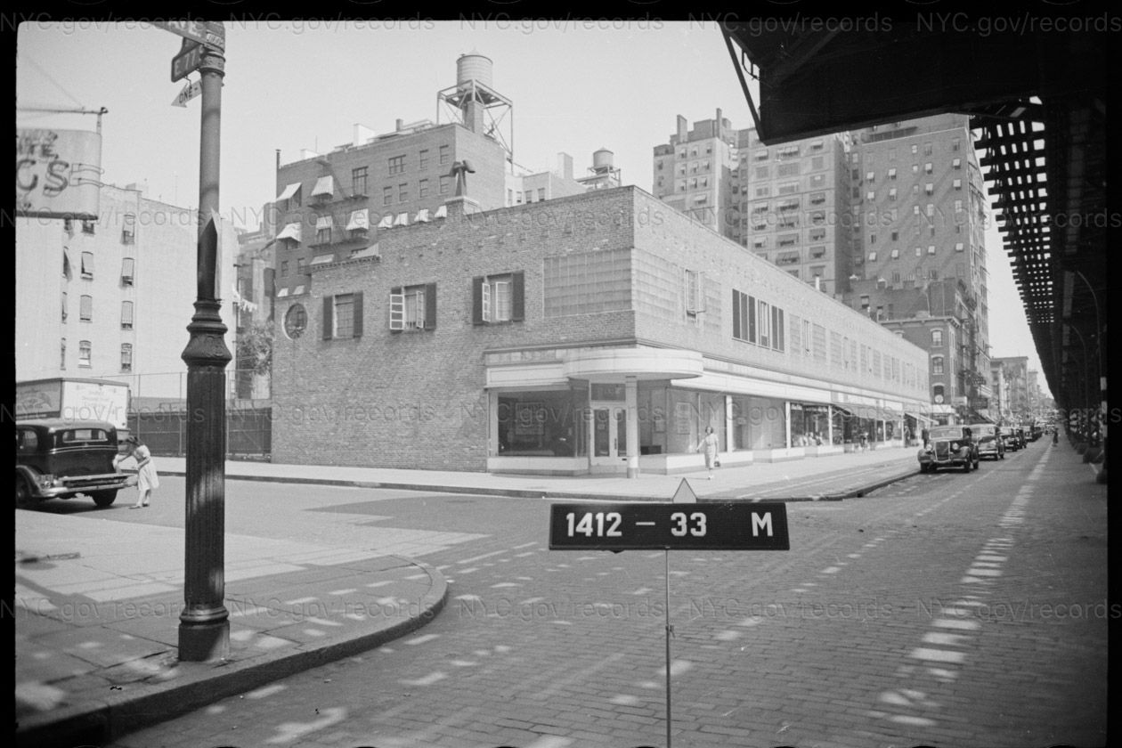 Tax photograph of a two-story corner building on Third Avenue: plate-glass shopfronts under a projecting rounded canopy, a pale banded frieze above them, and small windows in the brick second story. Its blind end wall on 77th Street has a round window and shuttered openings. The lattice shadow of the elevated railway falls across the roadway in the foreground.
