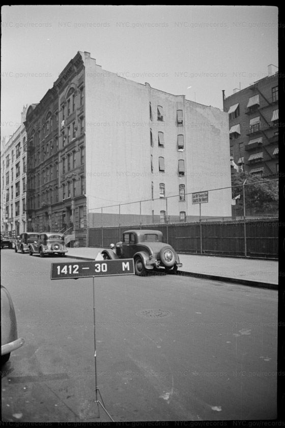 Tax photograph looking across East 77th Street at a chain-link fence backed by dark windscreen, enclosing open ground. A tall blank party wall of about six stories rises behind it, with a small two-line sign fixed to the fence. Brownstones with stoops stand at the left; 1930s cars are parked at the kerb.