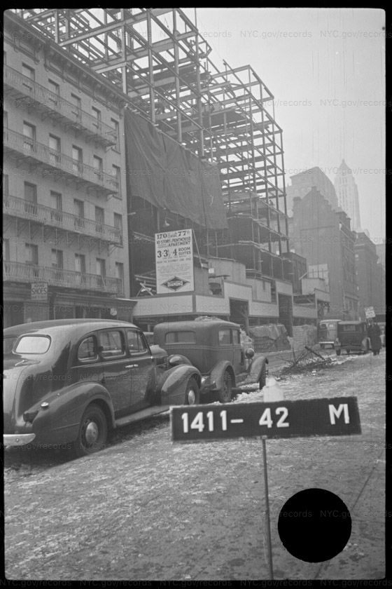 Winter tax photograph of a steel frame rising about ten stories behind a tarpaulin-draped hoarding, with an older apartment building and a shoe shop at the left, slush in the gutter, 1930s cars parked at the kerb and a tiered skyscraper visible in the distance.
