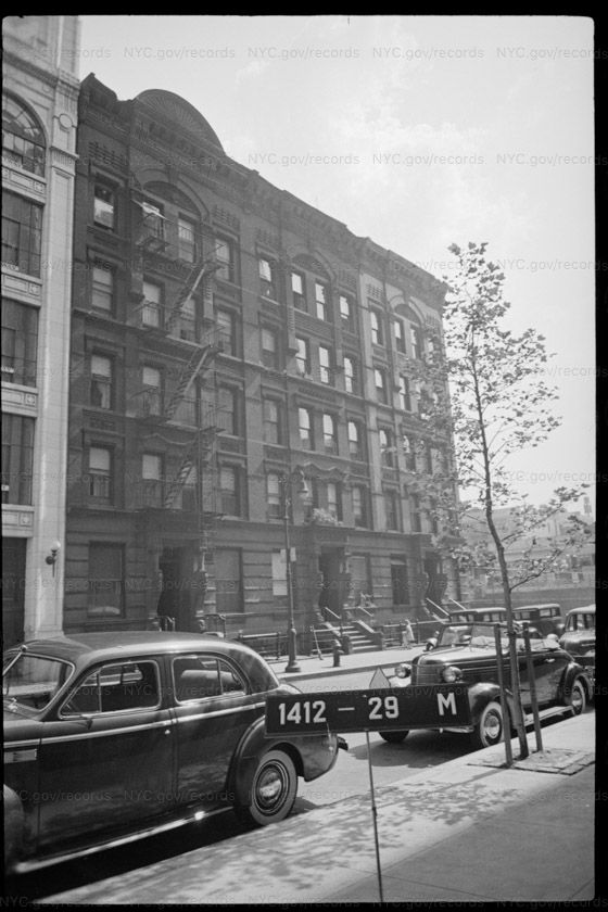 Tax photograph of a row of five-story brick tenements with iron fire escapes, arched window heads and high stoops, a newly planted street tree in front and 1930s cars at the kerb.