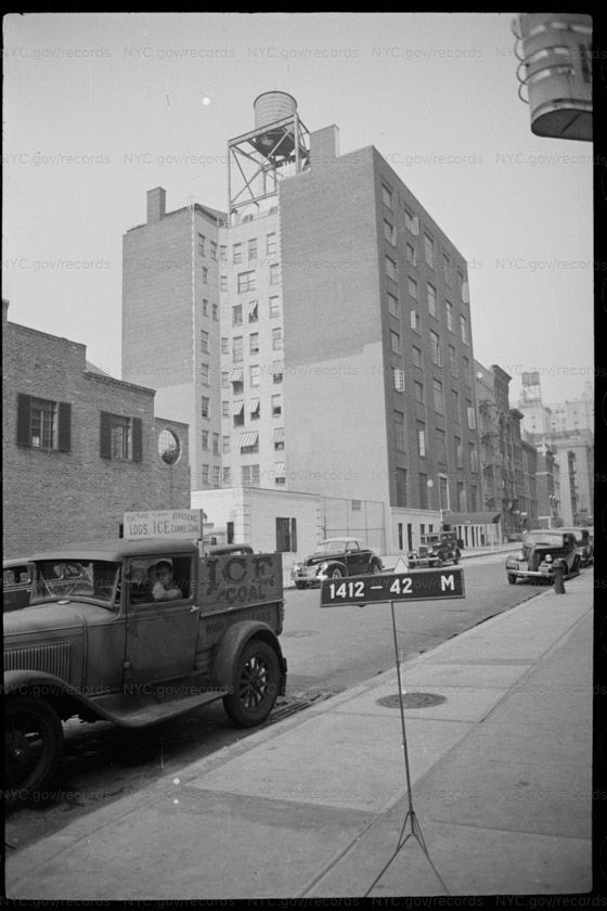 Tax photograph from East 78th Street looking into the middle of a block. A low brick building with shutters and a round window stands at the left, fenced open ground beyond it, and tall apartment houses with a rooftop water tank rise behind. An ice and coal dealer's truck is parked in the foreground, a man at the wheel.
