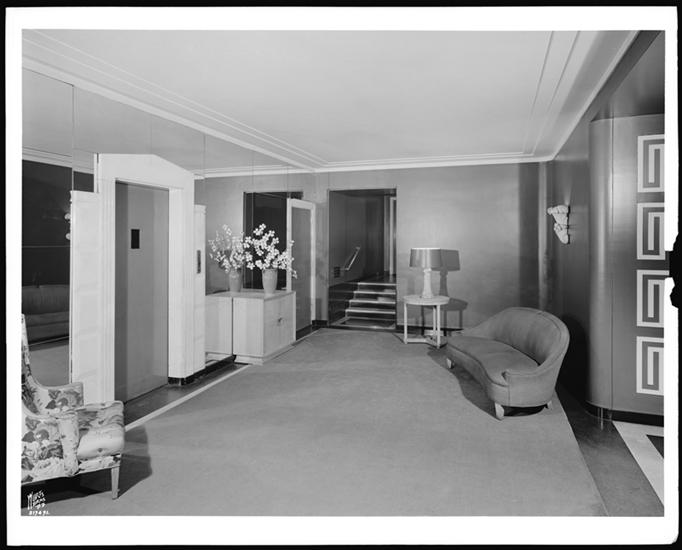 Architectural photograph of a Moderne apartment lobby: a stepped plaster cornice, mirrored wall panels flanking a pale elevator door, a blond wood cabinet with a vase of blossom, a shell-form wall sconce, a small round table with a lamp, a curved tub-shaped sofa on a plain fitted carpet, a few steps rising to a landing at the rear, and a curved wall at right incised with a repeating square-spiral pattern.