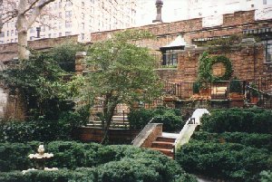 Colour snapshot of a walled interior-block garden with clipped box hedges in the foreground, a small stone ornament, and a flight of steps rising to a low brick building with a wreath on its wall.
